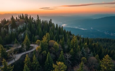 Wall Mural - Aerial view of a winding road through a dense forest on a mountainside, with a panoramic view of distant mountains and a lake at sunset.