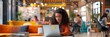 © nopommajun - A woman in an orange sweater is deeply engaged with her work on a laptop in a lively co-working space, portraying dedication and focus.
