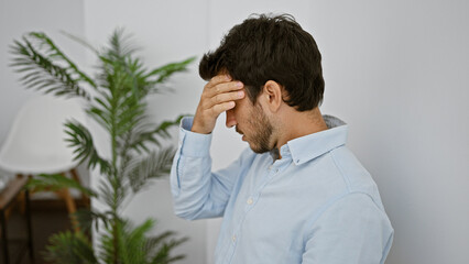 A young hispanic man in a blue shirt showing a sign of distress in a bright indoor setting.