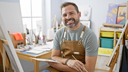 Handsome hispanic mature man with grey hair in a studio, wearing an apron and holding a paintbrush.