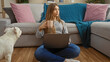 © Krakenimages.com - A young, beautiful blonde woman sitting with a laptop in a cozy living room while drinking from a mug, accompanied by her pet canine.