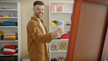 Wall Mural - Handsome bearded hispanic man taking a selfie in wardrobe room with smartphone, wearing glasses and casual clothes.