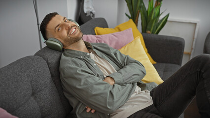 Wall Mural - A young hispanic man with a beard enjoys music on headphones, sitting relaxed in a cozy living room.