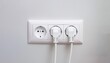 © Евгения Жигалкина - pristine white electrical plug is securely inserted into a standard electric socket mounted on a plain wall. The wall is smooth and unadorned, showcasing the simplicity and functionality of the plug