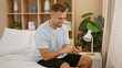 © Krakenimages.com - A smiling young hispanic man checks his smartphone while sitting in a modern bedroom with a cozy beige decor.