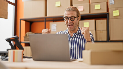 Excited man in glasses celebrating success at warehouse workplace with laptop and cardboard boxes.