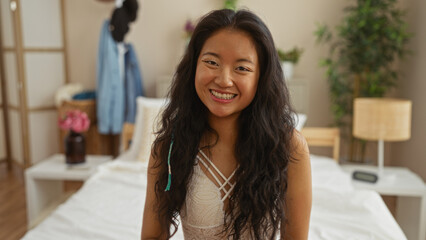 Beautiful young chinese woman smiling while sitting on a bed in a cozy bedroom in china with soft natural light and stylish decor.