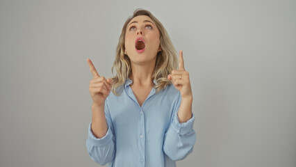 Poster - A surprised young woman with blonde hair pointing upwards, isolated against a white background, expressing astonishment.