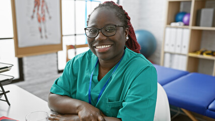 Canvas Print - African american woman with braids smiling in her workplace, a physiotherapy clinic interior, denoting professionalism and care.