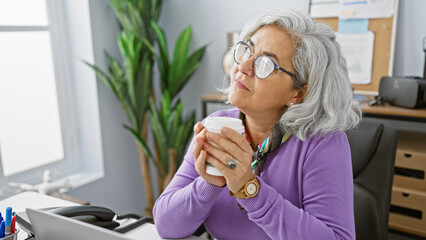 Poster - A thoughtful senior woman enjoys a coffee break at her modern office, displaying professionalism and tranquility.