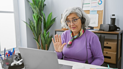 Sticker - A mature woman in glasses and a headset waves in a modern office setting, working on a laptop.