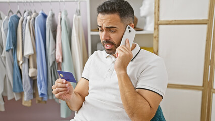 A concerned young hispanic man with a beard holds a credit card and talks on the phone in a dressing room.