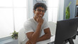 © Krakenimages.com - Young man smiling indoors in an office setting while sitting at a desk in front of a computer monitor, exuding a sense of happiness and productivity in a brightly lit room