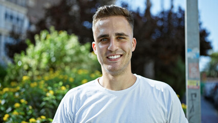 Poster - A smiling young hispanic man in a white shirt stands outdoors in a sunny park with greenery.