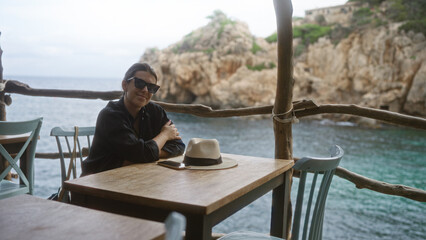 Poster - Young woman relaxing outdoors at cala deia in mallorca, spain, sitting at a wooden table with a view of the mediterranean sea and rocky cliffs in the background