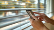 © Наталья Евтехова - An image of a person controlling smart blinds with a smartphone, with the blinds partially open and natural light streaming in, showcasing the seamless control of home features, wi