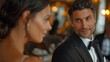 © Milan - Mature man attentively engaged in conversation with a woman at a formal event, dressed in a black tuxedo, exuding elegance and sophistication amidst softly lit surroundings.