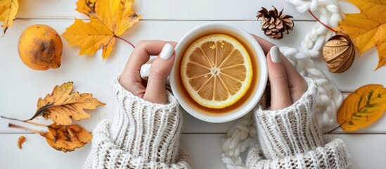 Poster - Woman in a cozy sweater holds a cup of tea with lemon against an autumn leaf background on a white table with room for a copy space image