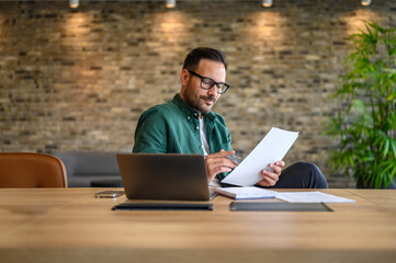 Wall Mural - focused male accountant in eyeglasses reading documents while working over laptop at desk in office