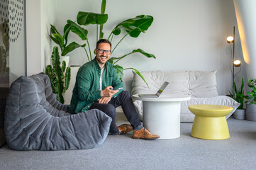 Wall Mural - Portrait of successful male entrepreneur with smart phone and laptop sitting on sofa in modern office