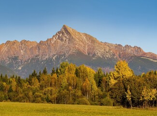 Poster - Beautiful autumn alpine landscape with a meadow, colored trees and an old wooden house, in the background a massive mountain ridge. Autumn walks in nature.