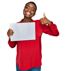 Wall Mural - Young african american woman holding blank empty banner smiling happy and positive, thumb up doing excellent and approval sign