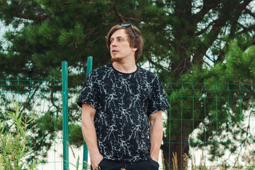 young man in casual attire standing outdoors near green trees during a sunny day