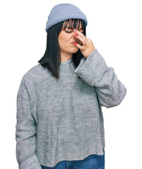 Poster - Young hispanic woman wearing cute wool cap smelling something stinky and disgusting, intolerable smell, holding breath with fingers on nose. bad smell