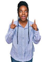 Sticker - Young african american woman wearing casual clothes amazed and surprised looking up and pointing with fingers and raised arms.