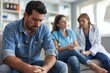 © Yuliia - male patient with a headache sitting on a couch during a therapy session at a clinic. The doctor and a woman psychiatrist are seen in the background, discussing treatment options.
