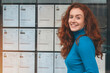 © Iryna - Young Woman Smiling in Blue Sweater Outside Information Board Displaying Announcements