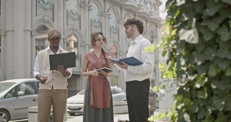 Canvas Print - Three business professionals engaged in a discussion while standing outside, using a laptop and notepads. The scene captures a collaborative and professional atmosphere.