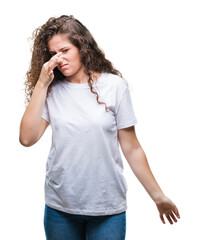 Beautiful brunette curly hair young girl wearing casual t-shirt over isolated background smelling something stinky and disgusting, intolerable smell, holding breath with fingers on nose