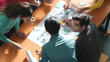 Top view of smart civil engineer working and writing blueprint while sitting at meeting table with blueprint, architectural equipment and colorful sticky note. Diverse team joining meeting. Symposium.