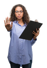 Wall Mural - Young hispanic business woman holding clipboard with open hand doing stop sign with serious and confident expression, defense gesture