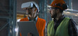 © Framestock - Two professional industry technicians wearing safety uniform and hardhats stand at manufacturing factory, use laptop and AR headset. Mature engineer and adult employee work in warehouse. Slow motion.