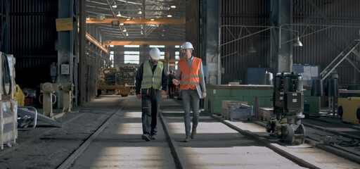 Two heavy industry engineers in safety uniform and hard hats discuss work process walking at manufacturing factory. Mature supervisor holds laptop, talks to adult worker in warehouse. Slow motion.