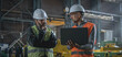 © Framestock - Two professional heavy industry workers, engineers wearing protective uniform and hardhats discuss work process using laptop computer while standing in manufacturing factory or warehouse. Slow motion.