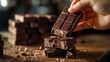 © Alex - A hand gently picks up a bar of dark chocolate surrounded by broken pieces, evoking warmth and indulgence in a cozy atmosphere