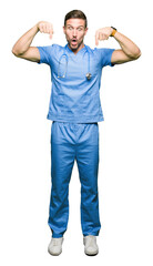 Sticker - Handsome doctor man wearing medical uniform over isolated background Pointing down with fingers showing advertisement, surprised face and open mouth