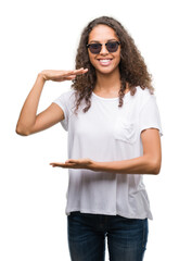 Wall Mural - Young hispanic woman wearing sunglasses gesturing with hands showing big and large size sign, measure symbol. Smiling looking at the camera. Measuring concept.