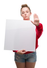 Canvas Print - Young blonde woman holding advertising banner with open hand doing stop sign with serious and confident expression, defense gesture