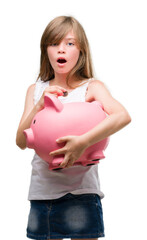 Poster - Young blonde toddler holding piggy bank scared in shock with a surprise face, afraid and excited with fear expression