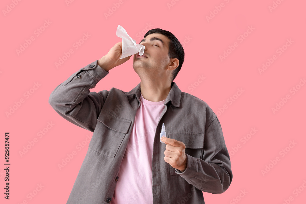 Ill young man with nasal drops and tissue on pink background
