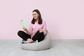 Poster - Young woman sitting on pouf and reading book near pink wall