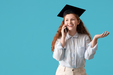 Wall Mural - Happy female student in graduation hat talking by mobile phone on blue background