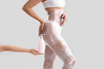 Poster - Young woman with stretch film and bottle of cream on light background. Body wrap treatment