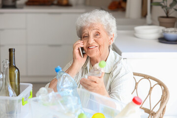 Wall Mural - Senior woman with containers of garbage talking by mobile phone at table in kitchen. Waste sorting concept