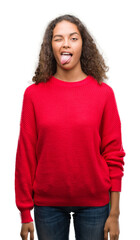 Wall Mural - Young hispanic woman wearing red sweater sticking tongue out happy with funny expression. Emotion concept.