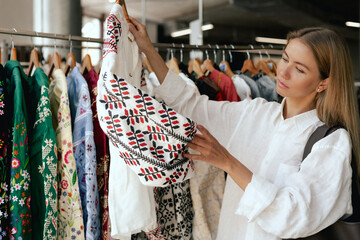 Middle aged woman runs across a rack of shirt with thread in a shopping mall. Ukrainian national clothes. Close-up of female hands plucked hanger choosing clothes in clothing store